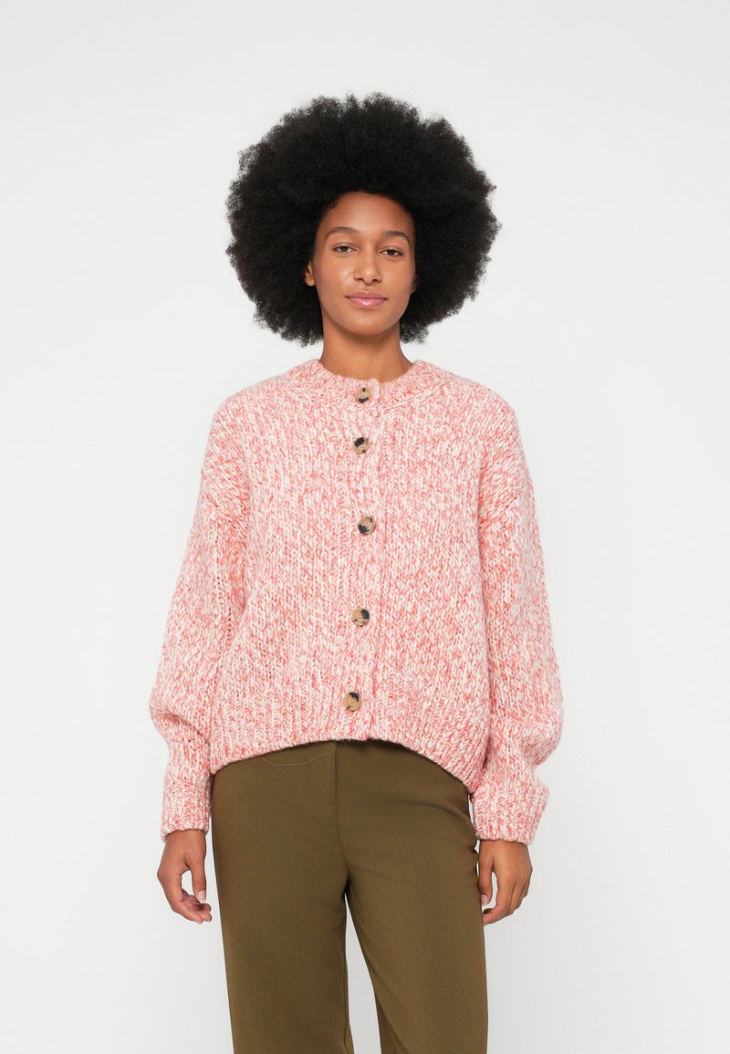 Mujer con cabello afro, vistiendo un cárdigan de punto rosa y blanco con botones y pantalones verde olivo, frente a un fondo blanco liso.