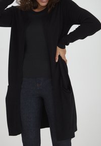 Zwarte lange cardigan met een open voorkant, geribbelde manchetten en een frontzak, gedragen met een donkere top en jeans. Soepele textuur, casual ontwerp.