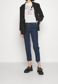 Blazer nero con dettagli a fibbia, t-shirt grigia con un cartone animato, jeans blu dritti e mocassini neri chunky.