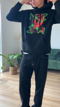 Schwarzer Hoodie mit einem rot-grünen Grafikdesign, ausgestattet mit einer Kängurutasche. Kombiniert mit schwarzen Hosen und in einem hellen Innenraum platziert.