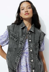 Veste en denim noir sans manches, avec des coutures blanches, des poches avant et des boutons argentés, superposée sur une robe à motifs bleus et blancs.