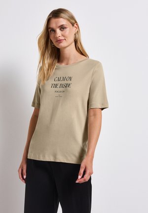 Camiseta de algodón beige con mangas cortas y escote redondo. En la parte frontal presenta una impresión negra: "CALMA POR DENTRO ENFOCA EN LA SERIE SILENCIOSA."