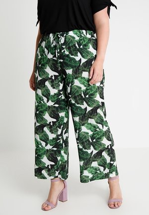 Kobieta w białych spodniach o szerokich nogawkach z zielonym tropikalnym liściastym wzorem i liliowymi sandałami na słupku stoi na jednolitym tle.