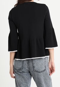 Černý pletený top s zvonovými rukávy a peplum spodním okrajem, který má bílé lemování na spodní části. Oblečeno s šedými džíny s vysokým pasem.