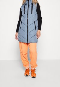 Frau in einer blauen gesteppte ärmellosen Jacke über einem schwarzen Langarmshirt, leuchtend orangefarbenen Jogginghosen und orangefarbenen Turnschuhen.