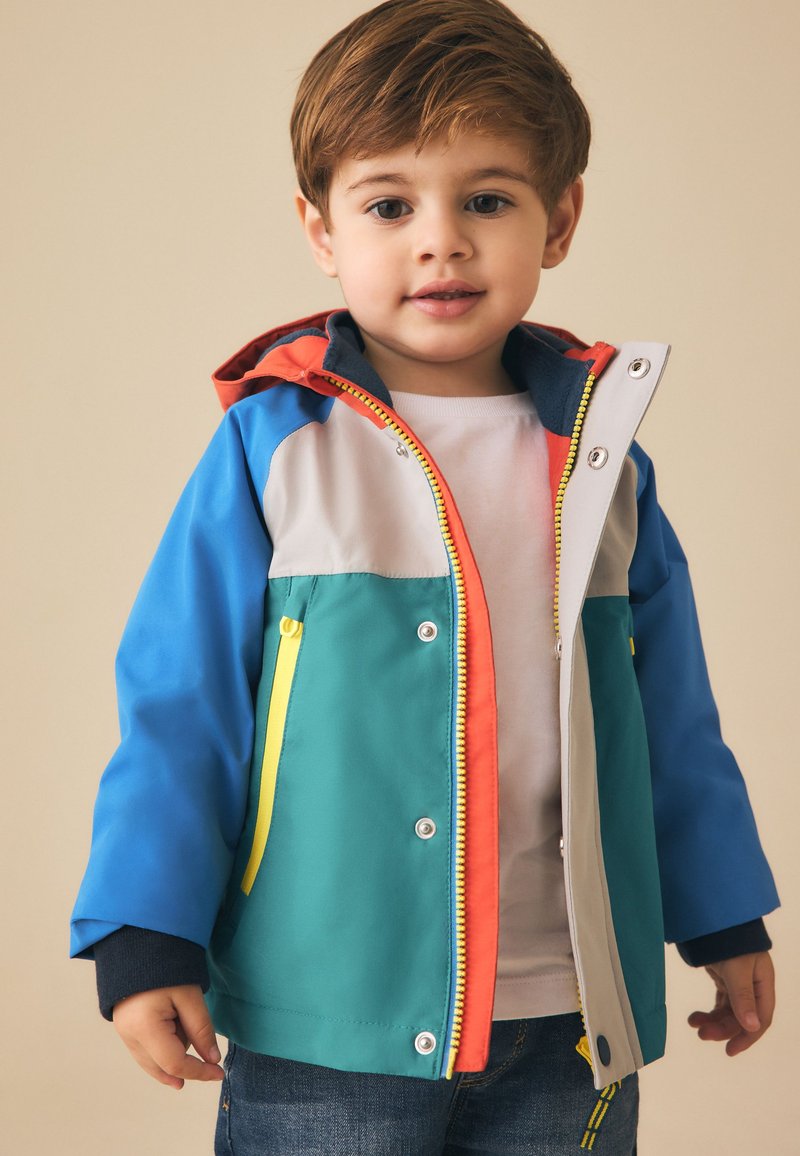 Jeune enfant portant une veste à capuche colorée avec des manches bleues, un devant bleu sarcelle, une poche zippée jaune et une doublure orange, debout devant un fond beige.