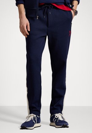 Pantalones jogger en azul marino de tejido suave con cintura ajustable, que cuentan con franjas laterales en contraste y un pequeño logo rojo en la pierna izquierda.