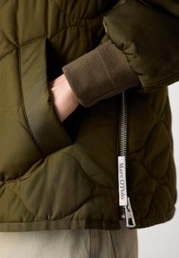 Main dans la poche d'une veste matelassée vert olive avec fermeture éclair visible et étiquette Marc O'Polo, manche en tricot avec bord-côtes et pantalon clair.