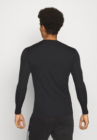 Muž s kudrnatými vlasy, oblečený v přiléhavém černém tričku s dlouhým rukávem a černých kalhotách, stojící zády k fotoaparátu před jednoduchým pozadím.