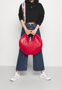 Sac à main rouge semi-circulaire en matériau lisse, avec une poignée découpée. Porté par une personne portant un sweat à capuche court noir et un jean à jambes larges.