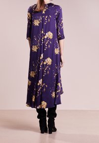 Długa fioletowa sukienka z żółtym wzorem kwiatowym, z rękawami do łokci i plisowanym tyłem, noszona na czarnych dopasowanych spodniach i botkach za kostkę.