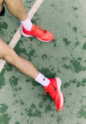 Scarpe da corsa Nike con una tomaia in mesh arancione brillante e swoosh nero, abbinate a calze bianche al polpaccio, su una superficie verde texturizzata.