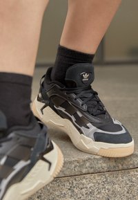 Black athletic shoes with a textured upper, featuring mesh and leather, contrasted with a light beige rubber sole and Adidas logo on the tongue.