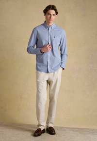 Jeune homme portant une chemise à carreaux bleue, un pantalon beige et des mocassins marron, debout devant un fond beige uni.