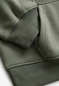 Sweatshirt vert olive avec des poignets texturés côtelés et un tissu lisse. Les coutures sont visibles le long des coutures et des bords.
