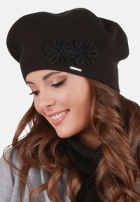 Černý pletený baret s volným střihem, zdobený dvěma tmavými květinovými aplikacemi a stříbrným akcentem. Měkká textura a zaoblený tvar.