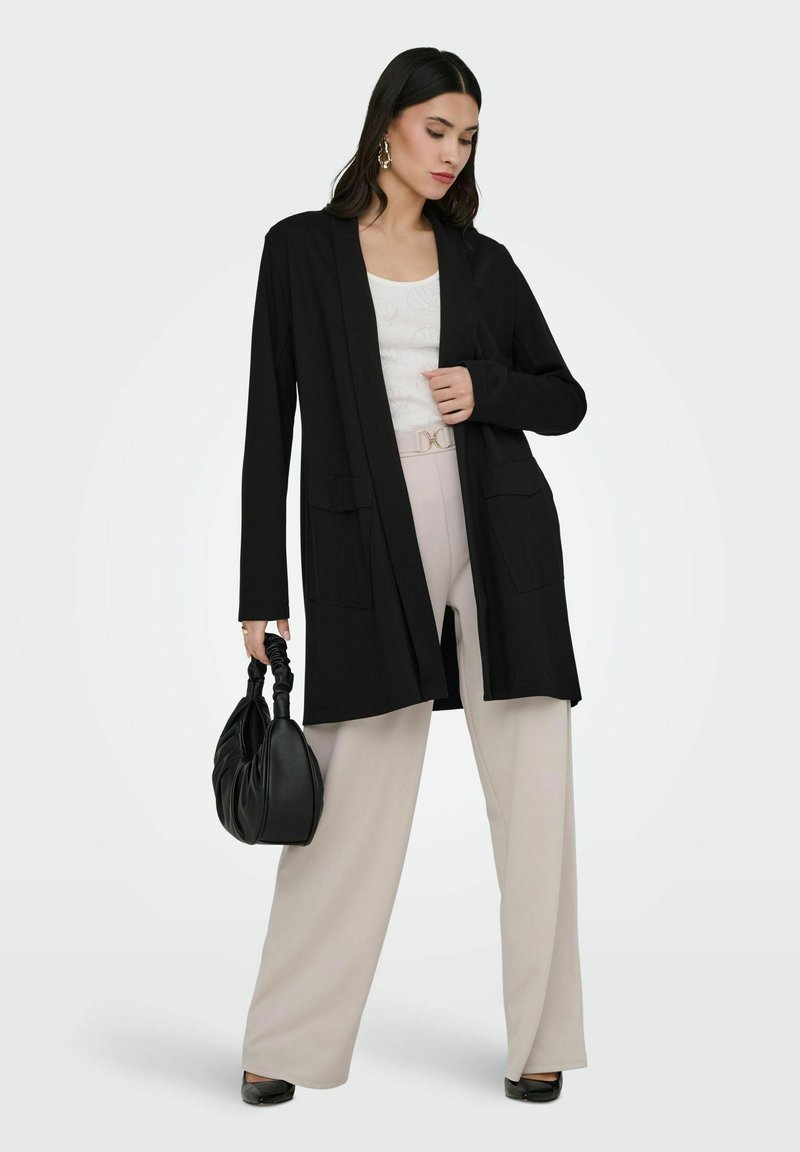Donna che indossa un lungo cardigan nero, top bianco, pantaloni beige a gamba larga, scarpe nere, che tiene una borsa nera con manico arricciato.