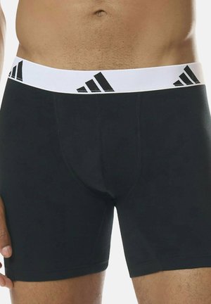 Närbild av en mans överkropp i svarta boxershorts med vit midja som har svarta Adidas-loggor.