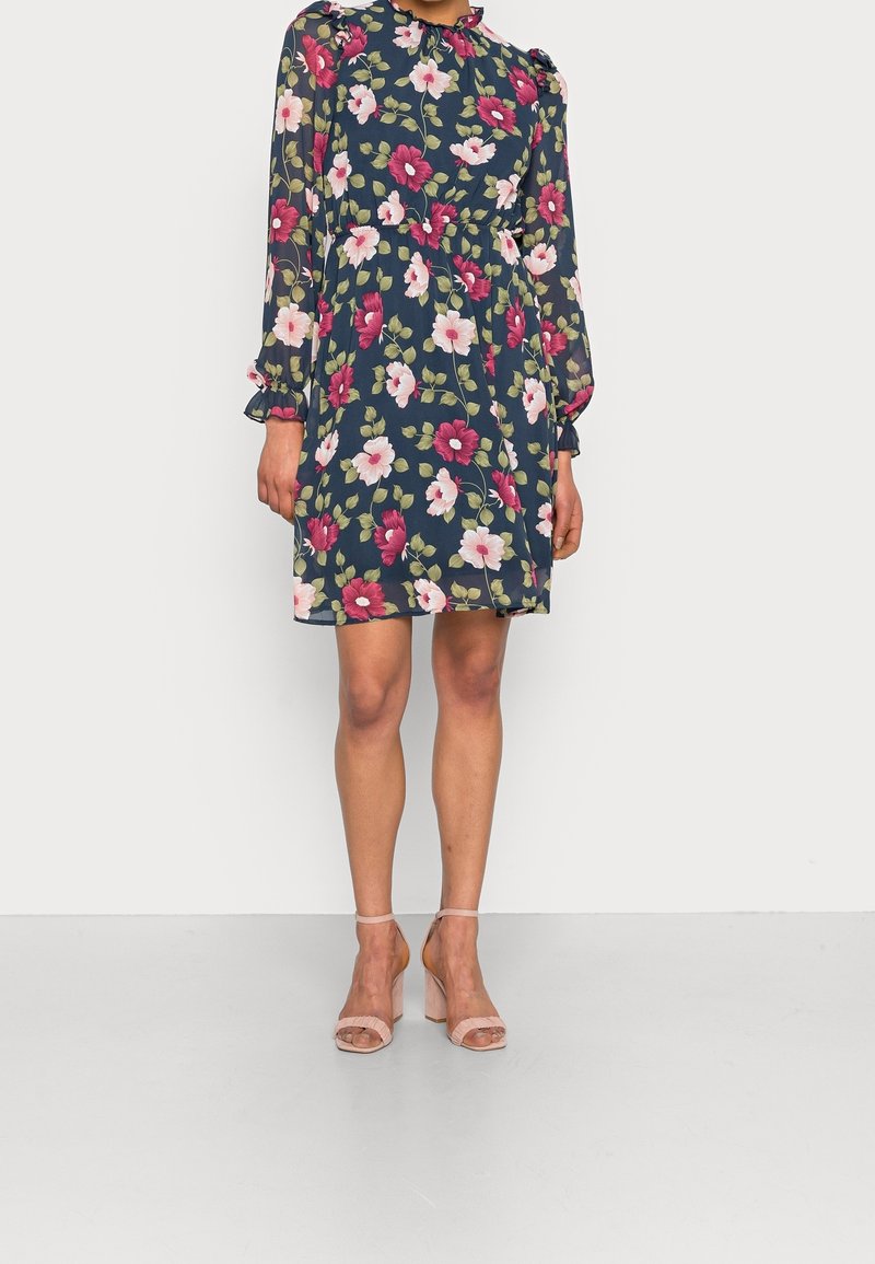 Femme portant une robe bleue marine à motifs floraux avec manches longues, col à volants, et sandales à talons blocs roses à bout ouvert, debout sur un sol gris.