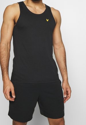 Muskuløs mand iført ensfarvet sort ærmeløs tanktop og sorte shorts, stående mod en neutral lys baggrund.