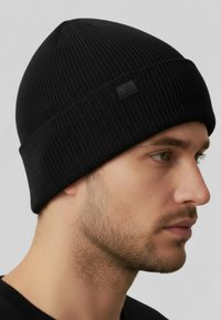 Man met lichte stoppels die een zwarte geribbelde gebreide beanie draagt, kijkend naar rechts tegen een neutrale achtergrond.