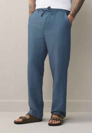 Mann trägt blaue Kordelhose und braune offene Sandalen, steht mit Händen in den Taschen vor neutralem Hintergrund.