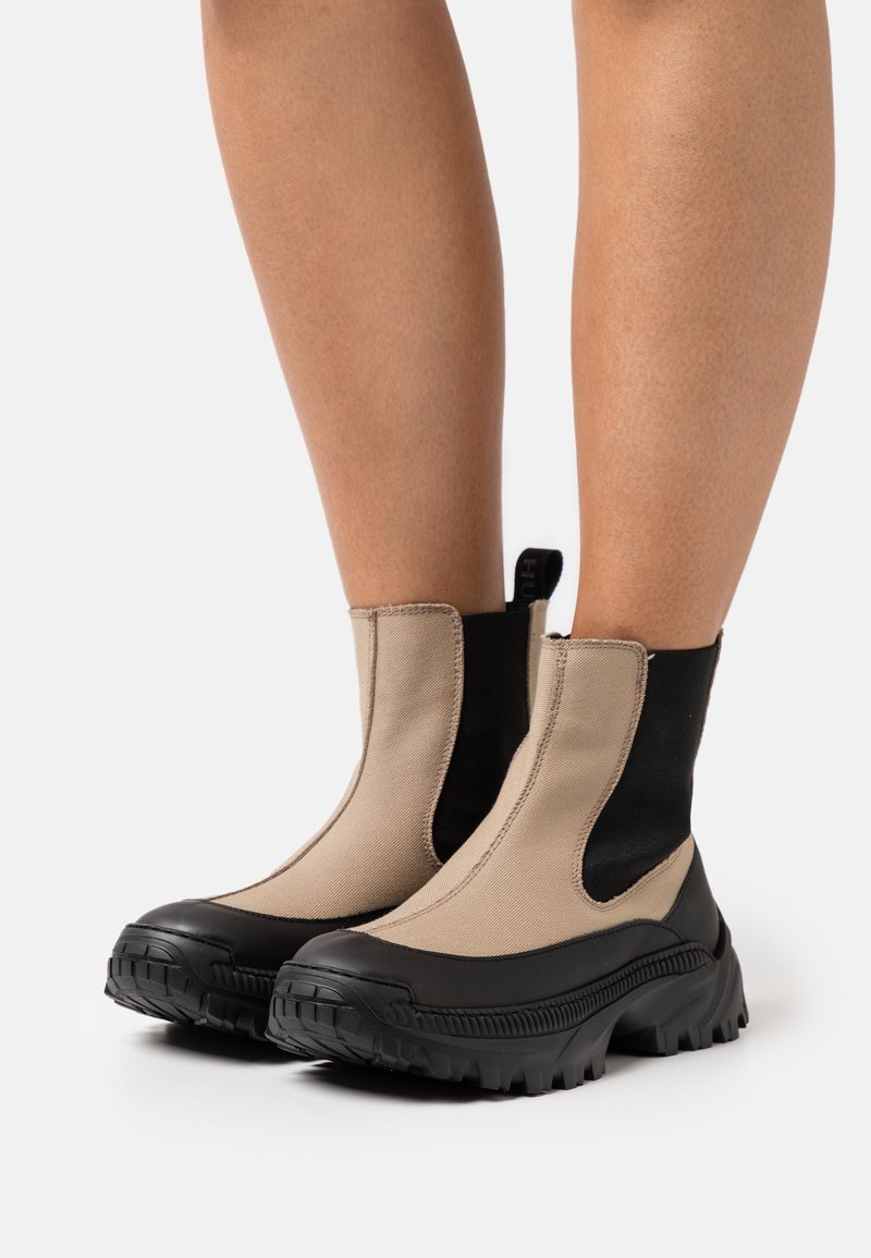 Bottines beige et noires avec semelles épaisses en caoutchouc portées par une personne, montrant les vues de face et de côté sur un fond uni.