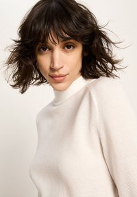 Frau mit kurzen, welligen dunklen Haaren, die einen hellbeigen strukturierten Rollkragenpullover trägt und direkt in die Kamera blickt, mit einem neutralen Hintergrund.