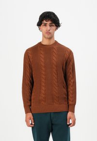 Giovane uomo con capelli ricci scuri indossa un maglione a trecce marrone e pantaloni verde scuro, in piedi davanti a uno sfondo bianco semplice.