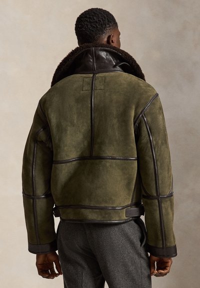 Mann trägt eine olivgrüne Wildlederjacke mit dunkelbraunem Lederbesatz und Kragen, steht mit dem Rücken zur Kamera.
