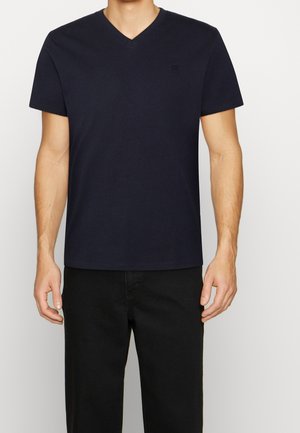 Homme portant un T-shirt uni bleu marine foncé à col en V et un pantalon noir, debout devant un fond blanc.