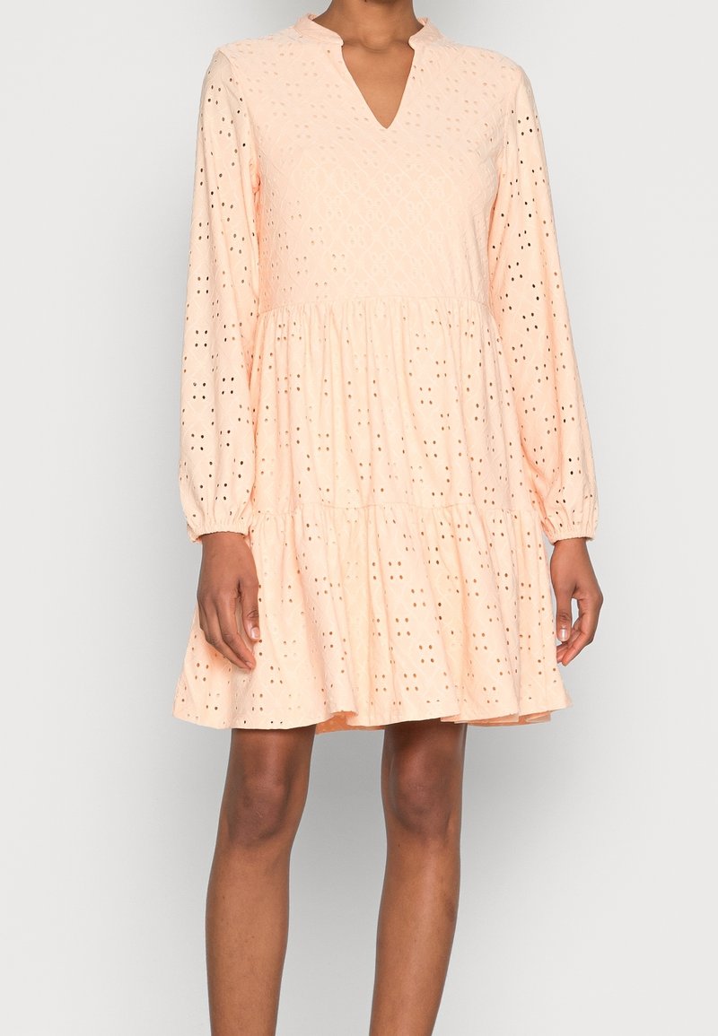 Femme portant une robe à manches longues couleur pêche avec un tissu ajouré et une jupe à volants, debout devant un fond uni.