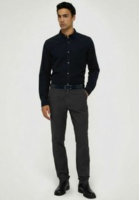 Tmavá košile na knoflíky s kapsou, spárovaná se šedými kalhotami a černými koženými botami, s úzkým střihem a hladkou texturou.