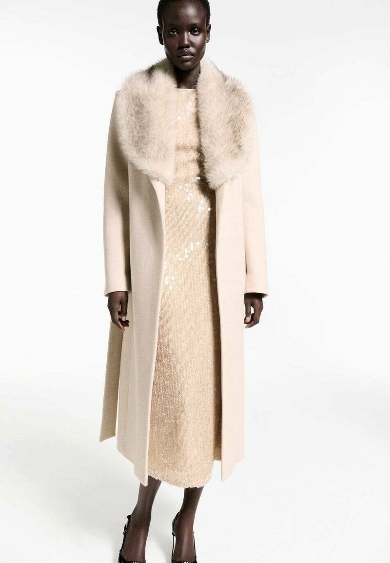 Beige wool coat with a faux fur collar, layered over a sparkly gold dress. The coat features long sleeves and a straight-cut silhouette.