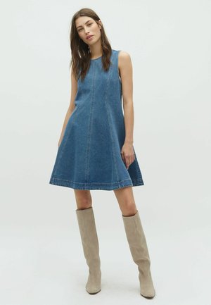 Femeie purtând o rochie albastră din denim fără mâneci și cizme bej din piele întoarsă până la genunchi, stând pe un fundal simplu și deschis la culoare.