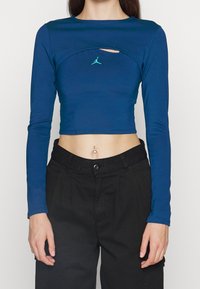 Modrý dlouhý rukáv crop top vyrobený z elastického materiálu, s kulatým výstřihem, výřezem na hrudi a malým logem ve světle zelené barvě.