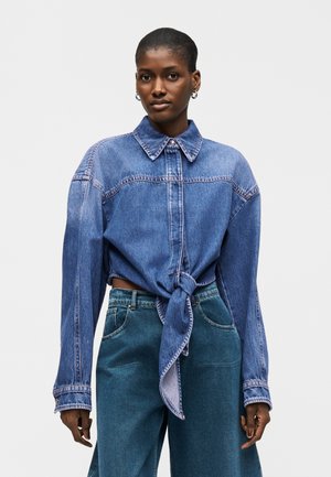 Γυναίκα με κοντά μαλλιά που φοράει κρίκους για σκουλαρίκια, μπλε τζιν πουκάμισο δεμένο και ψηλόμεσα μπλε τζιν παντελόνια σε απλό φόντο.