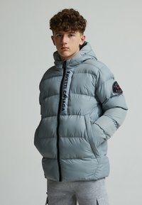 Veste matelassée gris clair avec un motif à coutures horizontales, capuche, fermeture éclair noire et accents de logo sur les manches, conçue pour la chaleur et le confort.