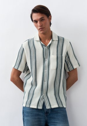 Νεαρός άνδρας με κοντομάνικο κρεμ πουκάμισο με κάθετες πράσινες ρίγες και τσέπη στο μπροστινό μέρος, στέκεται μπροστά σε λευκό απλό φόντο με τα χέρια πίσω από την πλάτη.