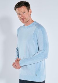 Hellblauer Strickpullover mit gerippten Details an den Ärmeln und Schultern, Rundhalsausschnitt und einem kleinen Logo an der unteren Seite.