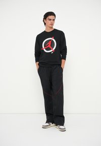 Μαύρη μακρυμάνικη μπλούζα με κόκκινη γραφική εκτύπωση και λευκές λεπτομέρειες, συνδυασμένη με μαύρο παντελόνι με κόκκινες ραφές και λογότυπο. Αθλητικά παπούτσια σε ανοιχτό γκρι και μαύρο.