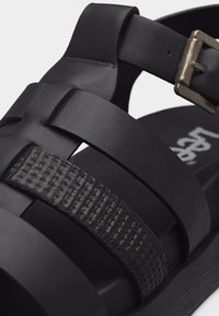 Μαύρο σανδάλι με υφασμένο λουράκι, λουριά από λείο δέρμα και μεταλλικό κούμπωμα σε επίπεδη σόλα.