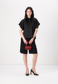 Camicia nera con fiocco, maniche a scallop, abbinata a pantaloni corti neri fino al ginocchio. Tiene una piccola borsa rossa. Tacchi alti neri.