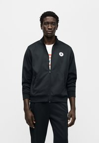 Μαύρο αθλητικό ζακέτο με φερμουάρ, με όρθιο γιακά, πλαϊνές τσέπες και λευκό κυκλικό λογότυπο στο στήθος. Φορεμένο πάνω από λευκό μπλουζάκι με γραφικό σχέδιο.