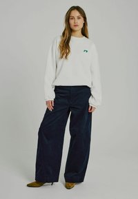Weißer Sweatshirt mit grünem Logo, kombiniert mit hoch taillierten, dunklen Cordhosen mit weitem Bein. Das Modell trägt braune spitze Absatzschuhe.