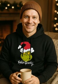 Felpa nera con una grafica vivace del Grinch con un cappello di Babbo Natale rosso. Il materiale sembra morbido e ha una vestibilità comoda. Persona che tiene una tazza beige.