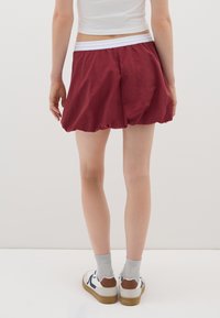 Persona che indossa pantaloni corti a bolle color marsala, una maglietta bianca, calzini grigi e sneakers bianche con tacco marrone, in piedi contro uno sfondo bianco semplice.