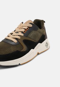 Baskets de sport pour hommes en vert olive, noir et beige avec tissu texturé, panneaux en daim, semelle blanche épaisse et lacets beiges.