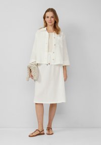 Witte, gestructureerde linnenjasje met knopen, gecombineerd met een bijpassende jurk. Een gehaakte tas en platte sandalen maken de look compleet.
