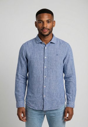 Junger Mann mit kurzen Haaren, der ein blaues Leinenhemd mit langen Ärmeln und Knöpfen sowie hellblaue Jeans trägt und vor einem schlichten grauen Hintergrund steht.